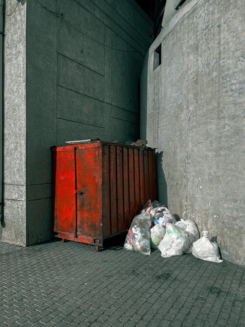 A large, rectangular metal refuse container with a rusty, weathered red finish, positioned against a dark green wall and adjacent to a light gray textured concrete wall. The container's surface shows signs of corrosion and chipped paint. Several white plastic bags filled with waste are piled next to the container on the paved ground, which consists of dark interlocking bricks. The setting appears to be an outdoor urban alley or backstreet area, with the container used for private rubbish disposal. In the background, there is a small, narrow window cut into the gray wall, and the lighting suggests daytime with natural light illuminating the space. This scene illustrates an example of an independent waste collection point, managed by local rubbish removal services, such as Rubbish Removal Haringey, supporting alternative methods of waste management outside of municipal rubbish collection.