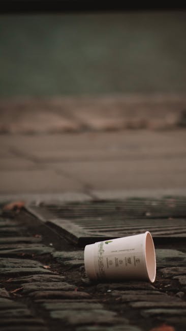 A white paper cup with printed text lies on a cobbled street near a metal drain cover, with the cup partially tilted onto its side. The background features a blurred section of a pavement or sidewalk, with subdued lighting suggesting an outdoor setting. The scene captures a small piece of roadside litter that may be encountered during street cleaning or private rubbish removal, illustrating the importance of proper waste disposal to prevent roadside debris. Rubbish Removal Haringey provides services aimed at maintaining clean, litter-free environments by removing such waste from external areas like streets and driveways, supporting local efforts to comply with waste management regulations and avoid fines. The focus on the cup and surrounding paving highlights typical roadside waste encountered in urban or suburban areas, emphasizing the need for efficient rubbish collection and site clearance to manage small-scale litter issues effectively and maintain a clean environment.