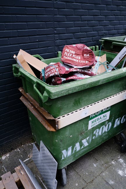 A green wheelie bin filled with black and red bags of garden waste, such as soil and mulch, placed on a paved surface beside a dark brick wall. The bin is positioned on top of a white plastic crate, and the bags display branding and product information. Behind the bin, there is a second, partially visible green bin. The scene suggests an area used for outdoor waste disposal or collection, with the bags likely prepared for a private rubbish removal service. The yellowish lighting and outdoor setting emphasize the practical and functional nature of waste management, aligning with services offered by companies like Rubbish Removal Haringey for alternative waste handling solutions besides municipal collection.