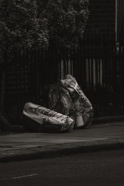 Three large black plastic rubbish bags, filled and tied at the top, rest on a paved sidewalk near a curb in an outdoor urban setting. The bags are slightly crumpled and appear to contain waste or unwanted items, with one of them partially leaning against a black metal fence that runs along the edge of a residential property. Behind the fence, dense foliage from a bush or tree is visible, creating a textured green backdrop. The area is shaded, with diffuse natural light casting mild shadows, indicating an overcast day or shaded environment. The scene is free of additional objects, emphasizing the presence of the waste bags as the primary element, which could be associated with private rubbish collection or alternative waste handling services, such as those offered by local rubbish removal companies including Rubbish Removal Haringey.