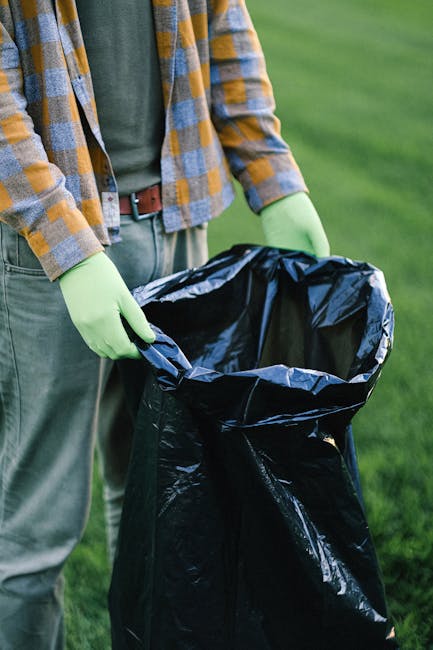 Three large black plastic rubbish bags, filled and tied at the top, rest on a paved sidewalk near a curb in an outdoor urban setting. The bags are slightly crumpled and appear to contain waste or unwanted items, with one of them partially leaning against a black metal fence that runs along the edge of a residential property. Behind the fence, dense foliage from a bush or tree is visible, creating a textured green backdrop. The area is shaded, with diffuse natural light casting mild shadows, indicating an overcast day or shaded environment. The scene is free of additional objects, emphasizing the presence of the waste bags as the primary element, which could be associated with private rubbish collection or alternative waste handling services, such as those offered by local rubbish removal companies including Rubbish Removal Haringey.