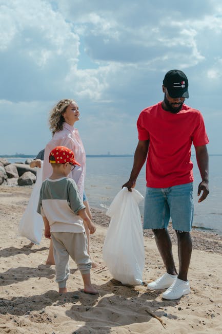 A young man dressed in a red t-shirt, light blue denim shorts, and a black cap with white and red details is standing on a sandy beach, holding a large white rubbish bag in his right hand while appearing to collect waste or debris. Behind him, a woman with curly blonde hair, wearing a light pink raincoat, observes the scene, standing near some rocks that form part of the shoreline. A child wearing a red and yellow cap, a light grey and white t-shirt, and beige pants is also present, facing away from the camera, holding another white rubbish bag. The background shows a body of water extending towards the horizon with a partly cloudy sky overhead, creating a natural outdoor setting that aligns with independent waste collection or environmental cleanup activities related to private rubbish removal efforts.