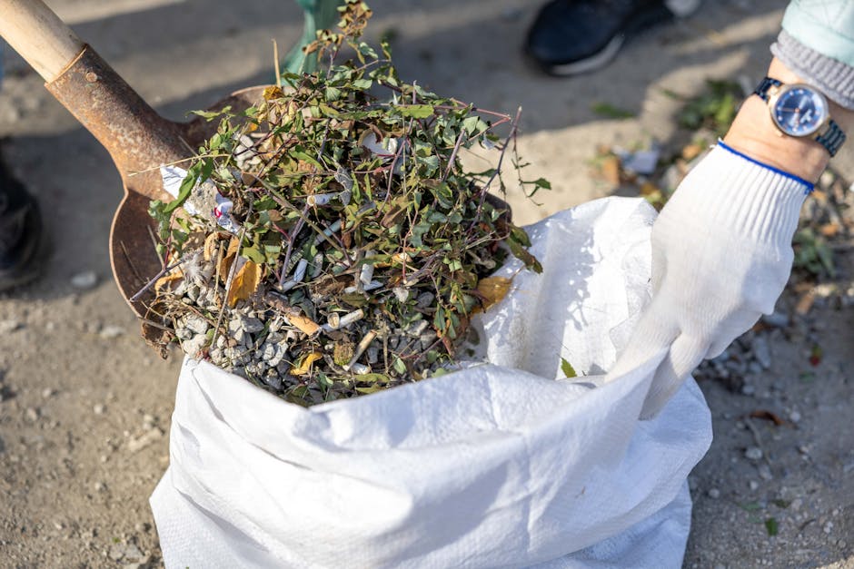 A person wearing a white glove and a watch is holding a white sack filled with various garden waste, including small branches, leaves, and gravel. The sack is open at the top, revealing the mixed contents, and is positioned on a gravel ground. In the background, part of a shovel with a rusted metal blade and wooden handle is visible, along with a small section of a person's black shoe. The scene suggests an outdoor area where garden clearance or waste collection is taking place, possibly as an alternative to traditional rubbish collection, with a focus on independent waste handling. The lighting is natural, casting soft shadows and highlighting the texture and colors of the garden debris and surrounding environment.