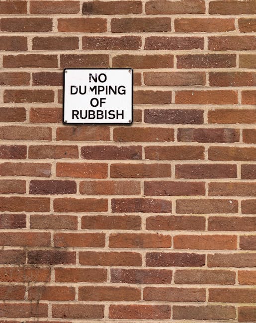 A close-up view of a red brick wall with a rectangular white metal sign mounted on it. The sign displays the text 'NO DUMPING OF RUBBISH' in black uppercase letters, with the word 'DUMPING' partially obscured or damaged, making part of it difficult to read clearly. The bricks are arranged in a horizontal running bond pattern, with some variation in the shades of red and brown, and visible mortar joints between them. The lighting is natural, casting soft shadows on the wall, and the overall scene suggests an outdoor environment, typical of areas where rubbish or waste disposal is restricted to prevent unauthorised dumping. This type of signage is often seen in locations managed by authorities such as the Haringey council or independent waste management services like Rubbish Removal Haringey to enforce waste disposal regulations and prevent illegal rubbish dumping.