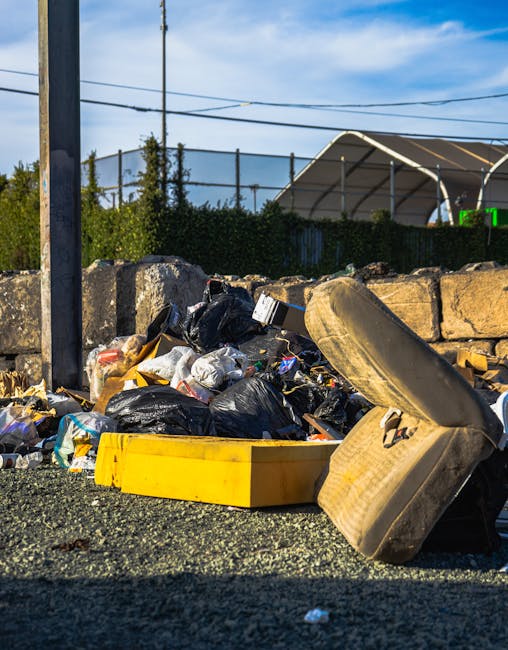 A pile of mixed household rubbish and waste materials is scattered on an outdoor surface, including black plastic bags, a yellow plastic tray, and an old, discarded beige car tyre lying on its side. The waste is positioned against a low stone wall constructed of roughly cut, uneven stones, with some debris spilling onto the gravel ground. In the background, there are green bushes and shrubs behind a metal fence, and a portion of a white, curved-roof outdoor structure or sports pavilion visible under a partly cloudy sky. A tall, vertical utility pole with overhead wires is situated on the left side of the scene, emphasizing an urban environment. The scene appears to be part of a private rubbish removal or disposal service, illustrating non-regular waste collection methods, aligning with the services offered by Rubbish Removal Haringey for alternative waste handling and bulky waste removal in the area.