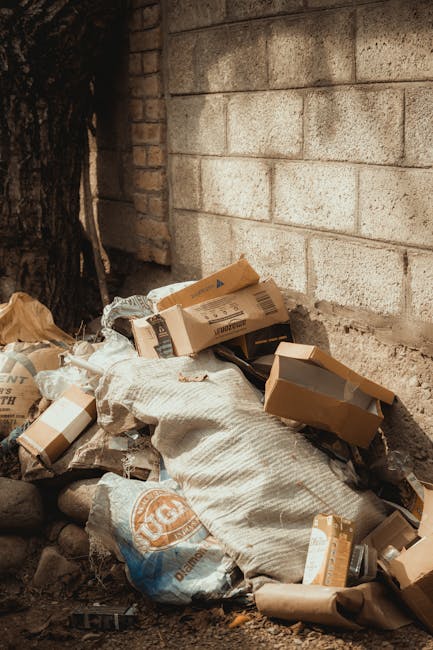 A pile of mixed household rubbish and waste materials is scattered on an outdoor surface, including black plastic bags, a yellow plastic tray, and an old, discarded beige car tyre lying on its side. The waste is positioned against a low stone wall constructed of roughly cut, uneven stones, with some debris spilling onto the gravel ground. In the background, there are green bushes and shrubs behind a metal fence, and a portion of a white, curved-roof outdoor structure or sports pavilion visible under a partly cloudy sky. A tall, vertical utility pole with overhead wires is situated on the left side of the scene, emphasizing an urban environment. The scene appears to be part of a private rubbish removal or disposal service, illustrating non-regular waste collection methods, aligning with the services offered by Rubbish Removal Haringey for alternative waste handling and bulky waste removal in the area.