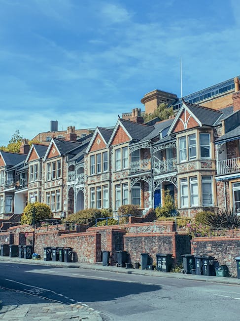 A row of Victorian-style terraced houses with ornate white decorative trim and bay windows, situated on an elevated brick terrace with small landscaped front gardens and low brick walls. The houses are constructed with red brick and feature slate roofs. In front, there is a paved street lined with multiple black wheelie bins arranged along the curb, indicating waste disposal activity. The sky above is clear and blue, with some wispy clouds, and the scene is lit by natural daylight, contributing to a bright and crisp atmosphere. The houses are adjacent to a modern building in the background, highlighting a contrast between historic and contemporary architecture, and suggesting the setting is in an urban residential area where private waste collection services, such as those offered by Rubbish Removal Haringey, might operate as an alternative to council rubbish collection.