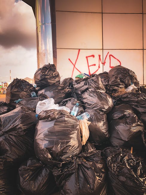 A large pile of black garbage bags filled with waste is stacked against a modern, tiled wall on an outdoor pavement section, with some white plastic bottles interspersed among the bags. The bags vary in size and appear tightly knotted at the top, with a few transparent sections revealing their contents. Behind the pile, the wall features graffiti with the word 'XEND' spray-painted in red. The scene is illuminated by natural light, with a cloudy sky visible in the background, suggesting a recent or ongoing rubbish collection or disposal activity that highlights the need for alternative waste handling options in the area. The presence of the bags and bottles indicates a typical street-level rubbish accumulation, possibly related to unscheduled or delayed rubbish removal services. Rubbish Removal Haringey's professional approach supports private or independent collection solutions, especially in cases of missed scheduled pickups or urgent waste clearance requirements, fitting within the broader context of non-local authority waste management.