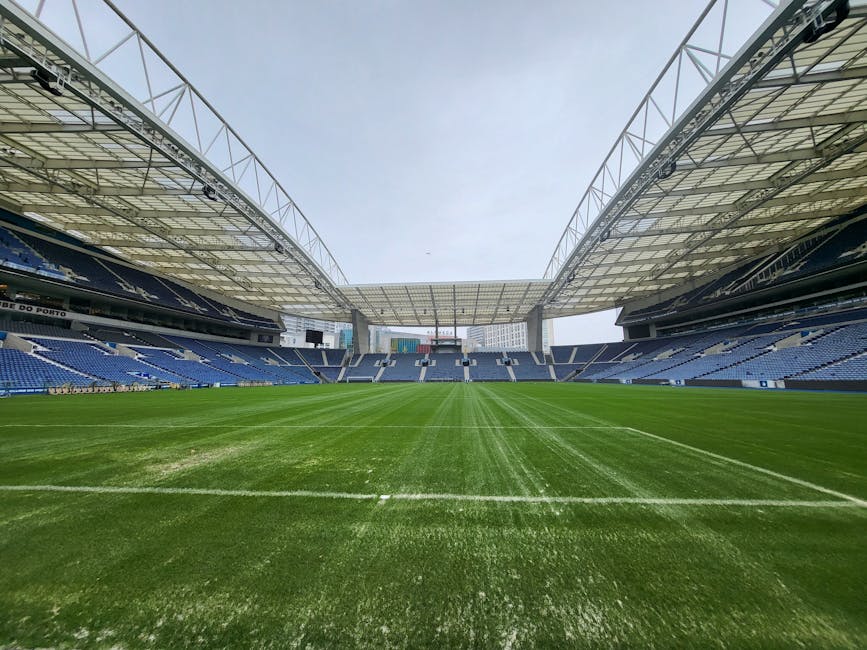 The image shows the interior of a large sports stadium with a well-maintained grass field in the center, surrounded by tiered seating areas with blue and white seats. The seating is arranged in multiple levels, with some sections accessible via staircases and walkways. Overhead, a modern, semi-transparent roof structure made of steel and glass panels extends over most of the seating, supported by steel trusses visible on either side. The sky above is clear and slightly overcast, providing diffuse natural light that illuminates the overall scene evenly. The grass on the playing surface is lush, vibrant green, with visible mowing lines running lengthwise across the field, indicating recent maintenance. In the background, tall buildings are faintly visible beyond the stadium walls, suggesting an urban location. The image gives an impression of a clean, empty venue prepared for a sports event, with no fixtures, equipment, or personnel present, emphasizing the spacious and orderly environment typical of a professional sports arena.