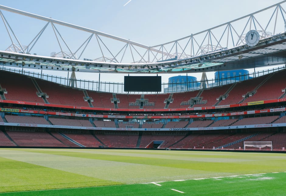 An expansive view of a football stadium interior during daylight shows a well-maintained grass pitch with alternating light and dark green stripes in the foreground. Surrounding the pitch are multiple tiers of red seating sections, with some grey seats interspersed. The stadium features a white, open lattice roof structure supported by tall, slender columns, allowing natural light to illuminate the area evenly. Visible digital scoreboards and advertising banners are positioned along the upper levels, with a few blue and black towers rising in the background outside the stadium. The overall environment appears clean and empty of spectators, illustrating a modern sports venue designed for large-scale events, which could relate to external or on-site waste management activities by companies performing auxiliary clearance or maintenance work within such facilities.