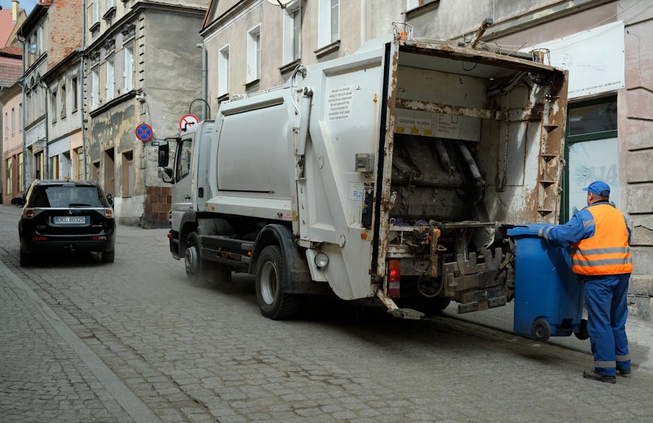A street scene showing a white rubbish collection truck operated by Rubbish Removal Haringey positioned with its rear door open, revealing mechanical lifting components used for waste compaction. The truck is parked on a cobblestone pavement alongside multi-storey residential buildings with stone and brick facades. To the right, a worker dressed in a blue uniform and orange high-visibility vest, wearing a blue cap and gloves, is seen emptying a blue wheeled bin into the truck’s rear compartment. A black car is parked further along the street’s left side, which is lined with other vehicles and limited traffic signs, including a no-parking and no-entry sign. The surroundings include aged building exteriors with some weathered paint and exposed brickwork, and the scene is lit with natural daylight, suggesting clear weather. This image visually captures the process associated with on-site waste collection services, illustrating the practical aspects of private rubbish removal operations in an urban environment while supporting the context of alternative waste handling methods.