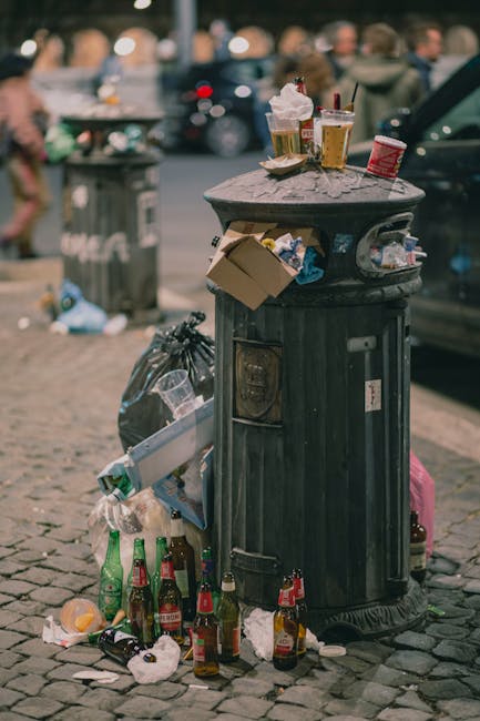 A street scene showing a white rubbish collection truck operated by Rubbish Removal Haringey positioned with its rear door open, revealing mechanical lifting components used for waste compaction. The truck is parked on a cobblestone pavement alongside multi-storey residential buildings with stone and brick facades. To the right, a worker dressed in a blue uniform and orange high-visibility vest, wearing a blue cap and gloves, is seen emptying a blue wheeled bin into the truck’s rear compartment. A black car is parked further along the street’s left side, which is lined with other vehicles and limited traffic signs, including a no-parking and no-entry sign. The surroundings include aged building exteriors with some weathered paint and exposed brickwork, and the scene is lit with natural daylight, suggesting clear weather. This image visually captures the process associated with on-site waste collection services, illustrating the practical aspects of private rubbish removal operations in an urban environment while supporting the context of alternative waste handling methods.
