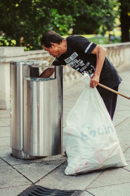 A middle-aged man with dark hair, wearing a black T-shirt with white graphic text, is seen in an outdoor urban environment during daylight. He is leaning forward slightly, holding a long-handled trash picker in his right hand, and is in the process of disposing of waste into a tall, shiny, stainless steel rubbish bin. The bin has a rounded, modern design with a vertical slit opening at the top. The man is gripping a large white plastic rubbish bag full of collected waste, which rests partially on the paved ground next to him. The surrounding environment features a stone balustrade in the background, and lush, green trees overhead, suggesting a park or a landscaped public area. The scene is well-lit by natural sunlight, highlighting the metallic finish of the bin and the textures of the pavement. This image exemplifies private waste disposal or rubbish collection in an outdoor public space, related to rubbish removal services offered by Rubbish Removal Haringey.
