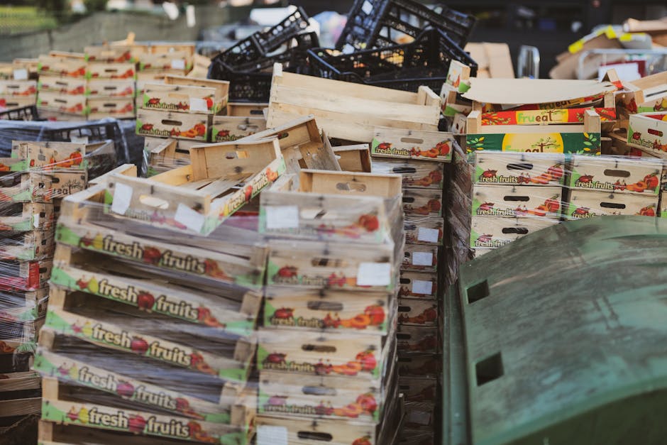 A large outdoor accumulation of cardboard produce boxes, predominantly labeled with 'fresh fruits' and colorful fruit graphics, stacked in multiple piles on a paved surface. The boxes are primarily light-colored with some showing signs of use, such as creases and slight wear, and are arranged in a haphazard manner, with some leaning or overlapping. Behind the stacks, there are a few black plastic crates and other discarded items, indicating an area designated for waste or recycling collection. To the right foreground, part of a green, weathered plastic container or bin is partially visible. The scene appears to be in an open container or storage yard, potentially related to a business or market waste collection operated by Rubbish Removal Haringey, specializing in private rubbish and waste removal services, with natural daylight illuminating the scene. The overall environment suggests a typical commercial waste sorting or disposal area, focused on refuse related to fresh produce packaging, ready for collection or disposal through an independent rubbish removal service.