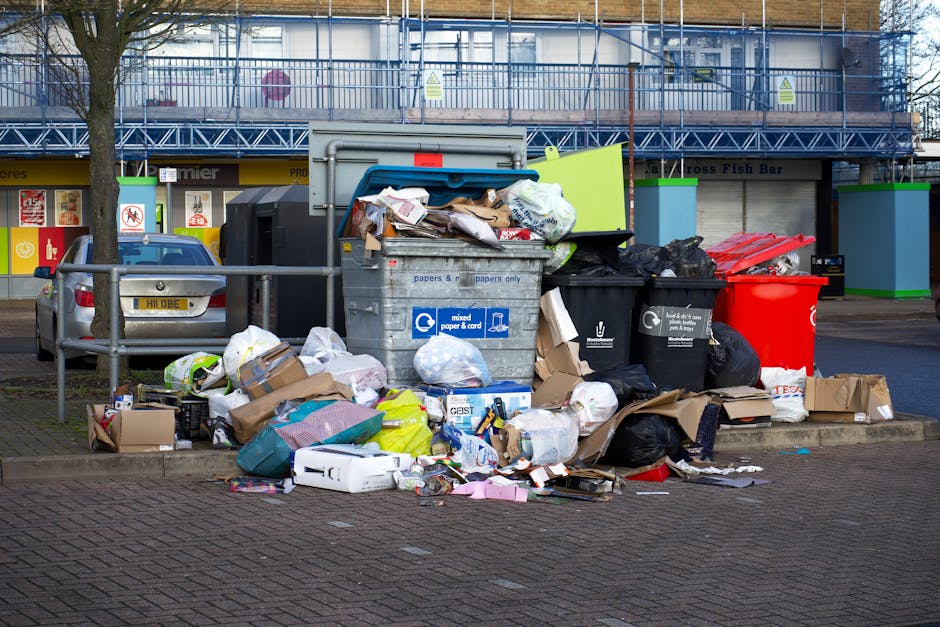 A middle-aged man with dark hair, wearing a black T-shirt with white graphic text, is seen in an outdoor urban environment during daylight. He is leaning forward slightly, holding a long-handled trash picker in his right hand, and is in the process of disposing of waste into a tall, shiny, stainless steel rubbish bin. The bin has a rounded, modern design with a vertical slit opening at the top. The man is gripping a large white plastic rubbish bag full of collected waste, which rests partially on the paved ground next to him. The surrounding environment features a stone balustrade in the background, and lush, green trees overhead, suggesting a park or a landscaped public area. The scene is well-lit by natural sunlight, highlighting the metallic finish of the bin and the textures of the pavement. This image exemplifies private waste disposal or rubbish collection in an outdoor public space, related to rubbish removal services offered by Rubbish Removal Haringey.